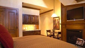 A bedroom with a large bed, a fireplace, a television mounted above it, and a dining area with a table and chairs.
