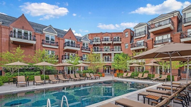 A poolside area with lounge chairs and umbrellas, a brick building with balconies, and a clear blue sky.