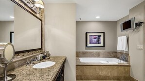 A bathroom with a double sink, a large mirror, a framed picture on the wall, a TV mounted above the bathtub, and a towel rack.
