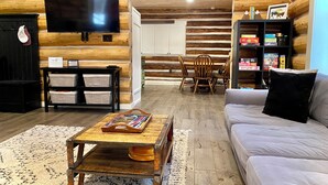 A log cabin interior with a TV, wooden furniture, a bookshelf, and a dining area. - Walk to Lake, Downtown, Restaurants and Shops- Grand Mountain Haven (Grand Lake)