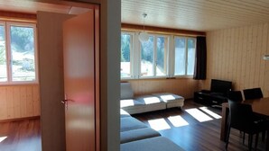 Sala de estar moderna com sofá, mesa de jantar e cadeiras. Janelas amplas permitem entrada de luz natural.
