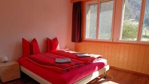 Quarto com piso de madeira, cama com roupa de cama vermelha, mesa de cabeceira e janela com cortinas.