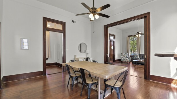 Un comedor con mesa y sillas de madera, ventilador de techo y vista a otra habitación con sofá y cortinas.