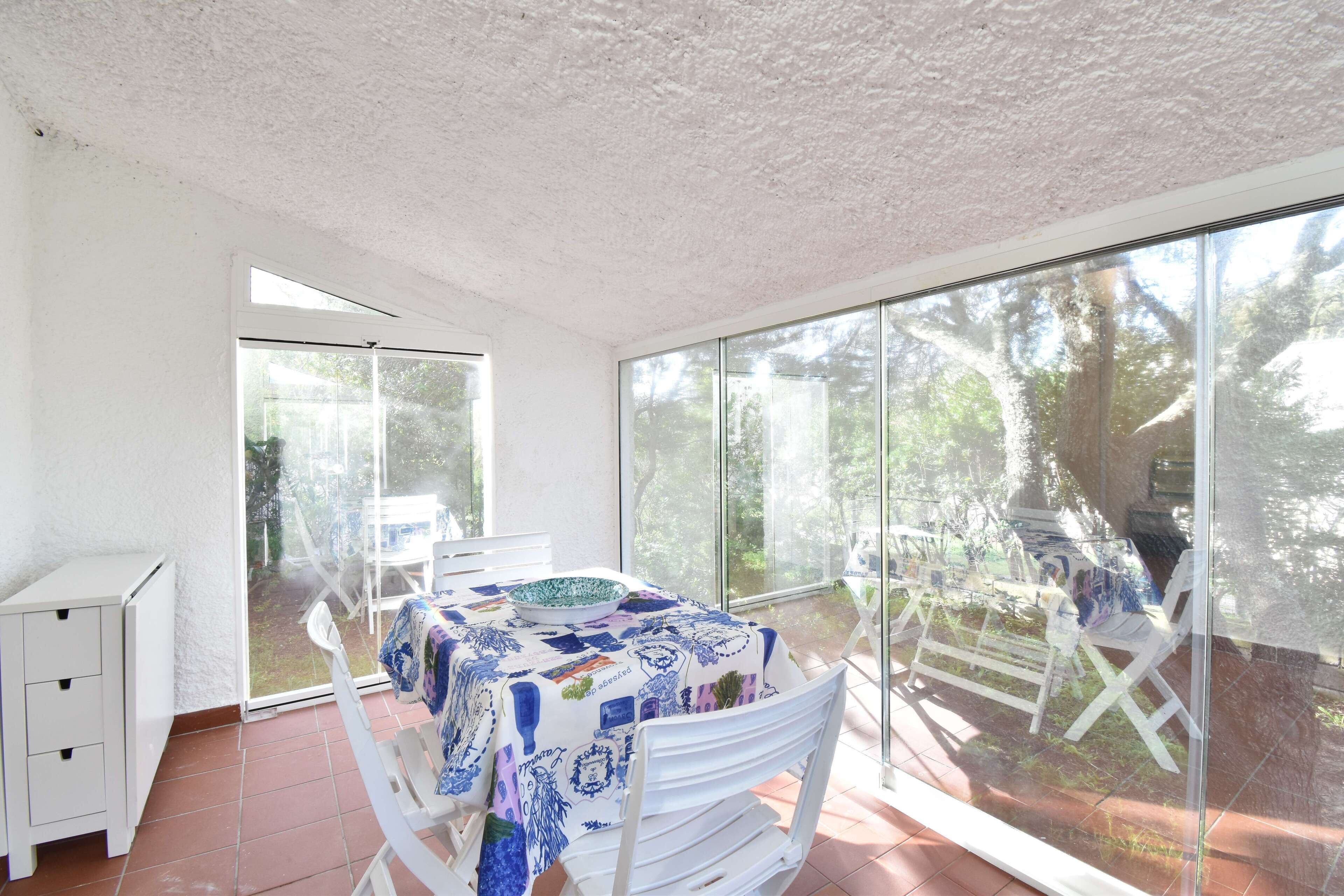A room with a large sliding glass door leading to a garden, a table with a patterned tablecloth, and a white chair.