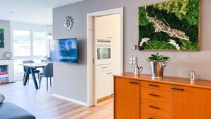 Ein modernes Wohnzimmer mit Flachbildfernseher, Essbereich mit Tisch und Stühlen, einer Holzkommode mit Schubladen und einem großen Wandkunstwerk aus grünem Moos.