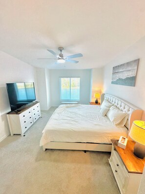Une chambre à coucher avec un lit, un téléviseur posé sur une commode, un ventilateur de plafond, une fenêtre et un tableau accroché au mur.