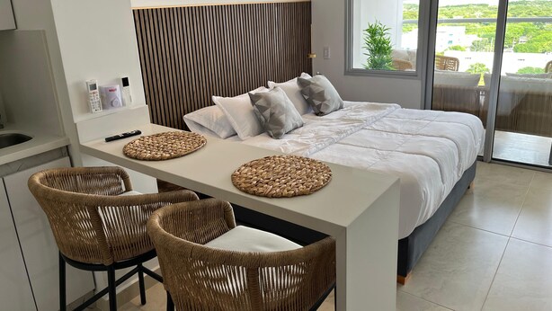 Un dormitorio moderno con una cama grande, una mesita pequeña, dos sillas de mimbre y vistas a la vegetación por la ventana.
