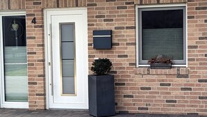 En murstensbygning med en hvid dør, vindueskasser og en postkasse. En lille plantekasse med blomster står foran.