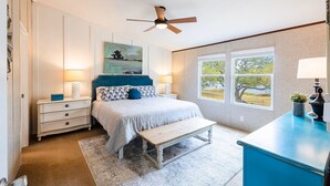Une chambre à coucher comprenant un lit, des tables de chevet, un banc, un ventilateur de plafond et une fenêtre donnant sur des arbres.
