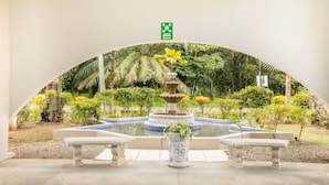 A fountain with a tiered design, surrounded by a garden with various plants and a bench in the foreground. - With preferential price for monkey tours (Quepos)
