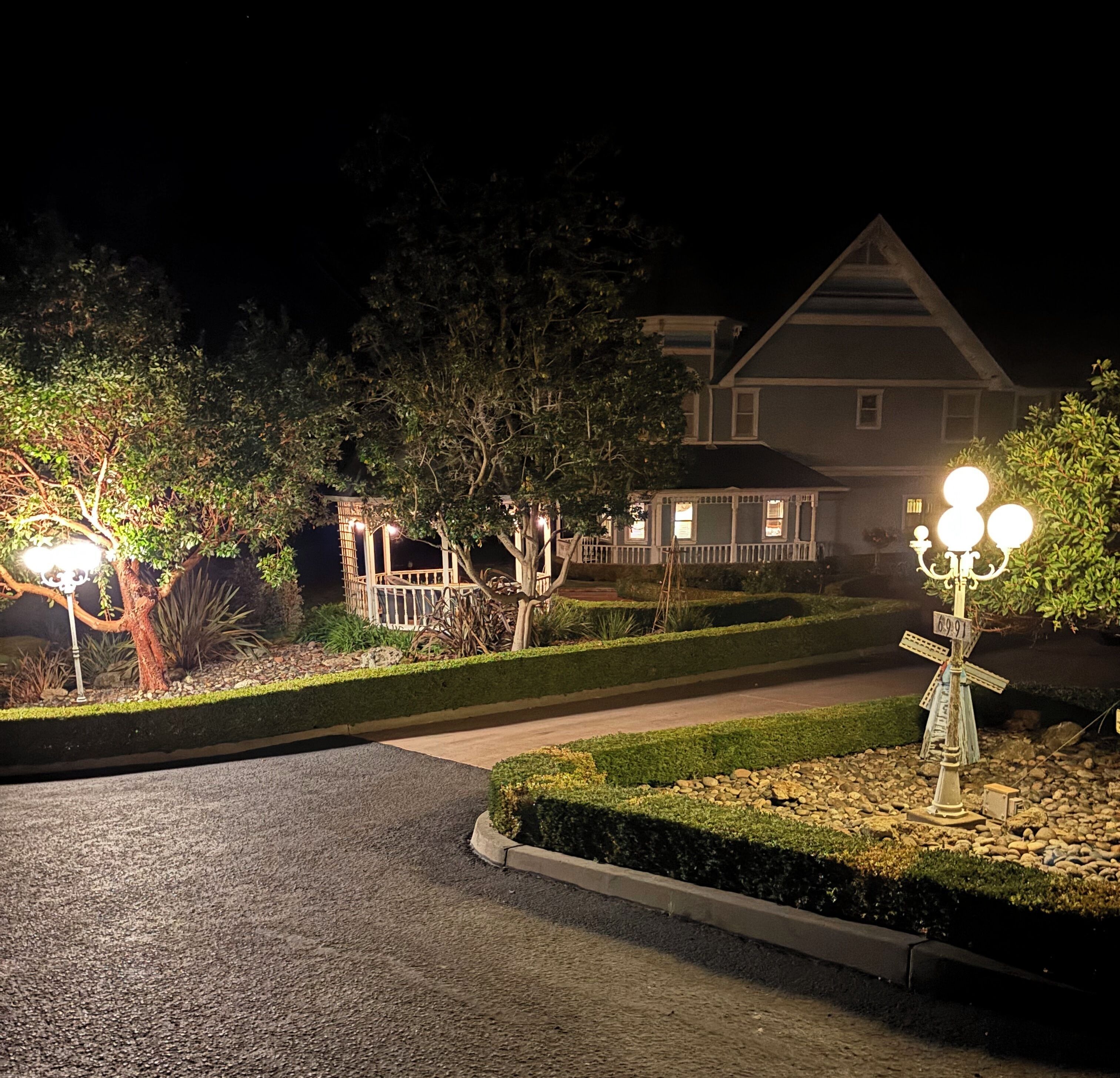 Une allée d’accès résidentielle bien éclairée la nuit, avec une maison en arrière-plan, entourée de jardins aménagés et d’arbres.