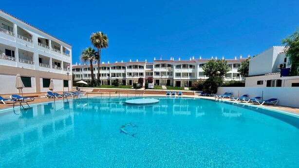 A clear blue swimming pool with lounge chairs and a palm tree, surrounded by a multi-story building with balconies. - Sa Caleta Apartment with swimming pool next to the beach (Cala En Blanes)