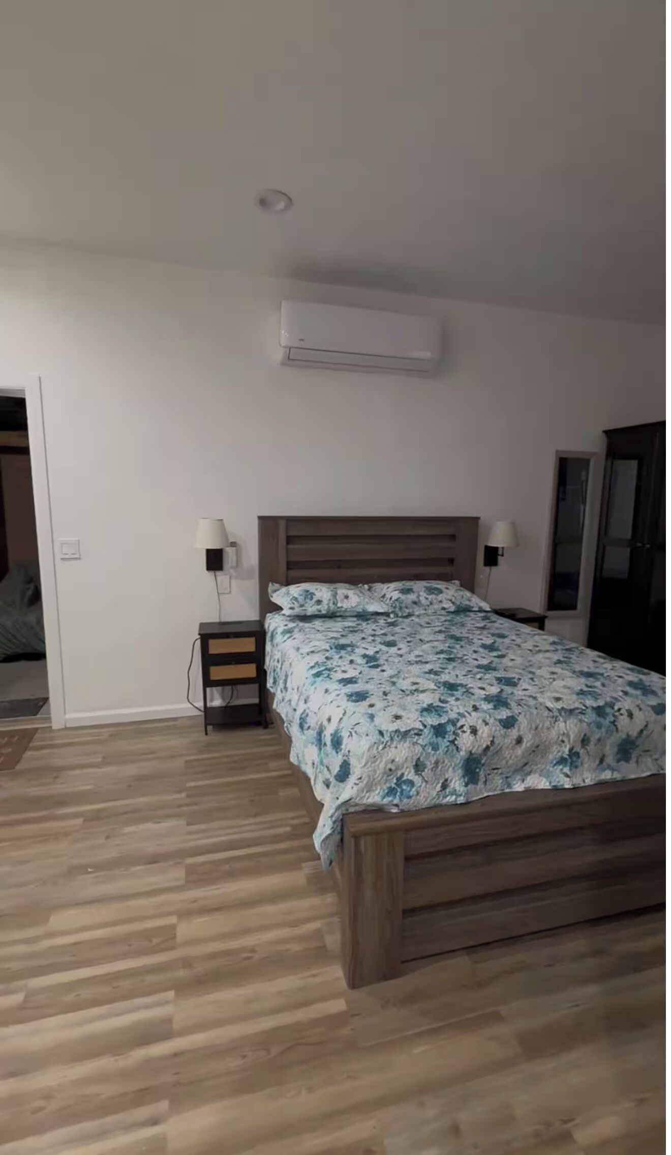 Ein Schlafzimmer mit einem hölzernen Bett, einem Nachttisch, einer Wand-Klimaanlage und einer Tür zu einem weiteren Raum.