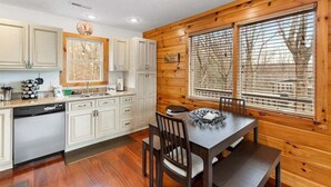 A kitchen with wooden walls, a dining table with chairs, and a clock on the wall. - Stunning Mountain Retreat! Packed with Fun! Air Hockey Table! - Two Sisters Chalet (Fancy Gap)