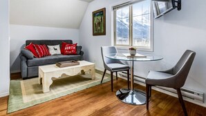 Un salon chaleureux avec un canapé gris, une table ronde en verre et deux fauteuils gris. Un téléviseur à écran plat est fixé au mur, au-dessus de la table.