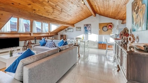 Ein großzügiger Wohnbereich im Loft-Stil mit hoher Decke und freiliegenden Holzbalken, ein großes graues Sofa mit blauen Kissen, ein Flachbildfernseher und ein Essbereich mit Tisch und Stühlen.