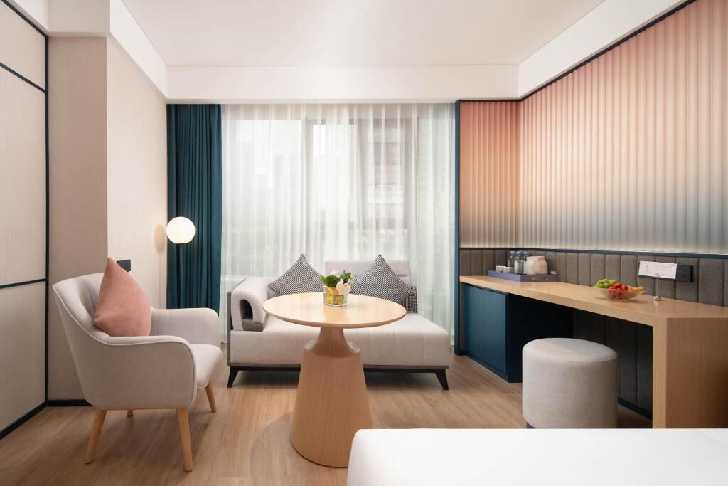 A modern hotel room with a sofa, armchair, round table, and a small kitchen area.