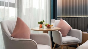 A modern hotel room with two armchairs, a small round table with a fruit bowl and a plant, and a view of the city through sheer curtains. - Guangzhou Chimelong Expo Center Jincheng Hotel (Guangzhou)