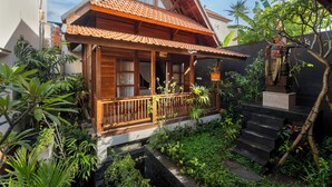 A wooden house with a tiled roof, a wooden porch, and a garden with various plants and a small staircase leading to the entrance. - KIRANI HOUSE (Batubulan)