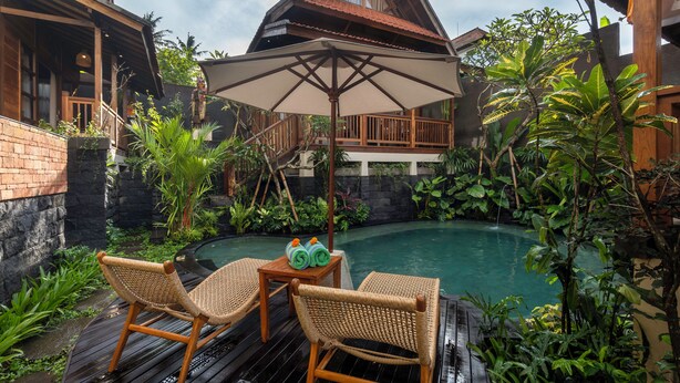 A wooden deck with two wicker chairs and a table, a pool, and lush tropical plants. - KIRANI HOUSE (Batubulan)