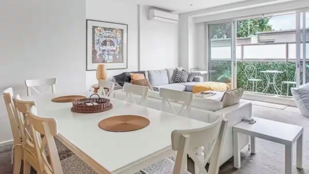 Ein modernes Wohnzimmer mit einem weißen Sofa, einem Esstisch und Stühlen. Ein großes Fenster bietet Blick ins Freie.