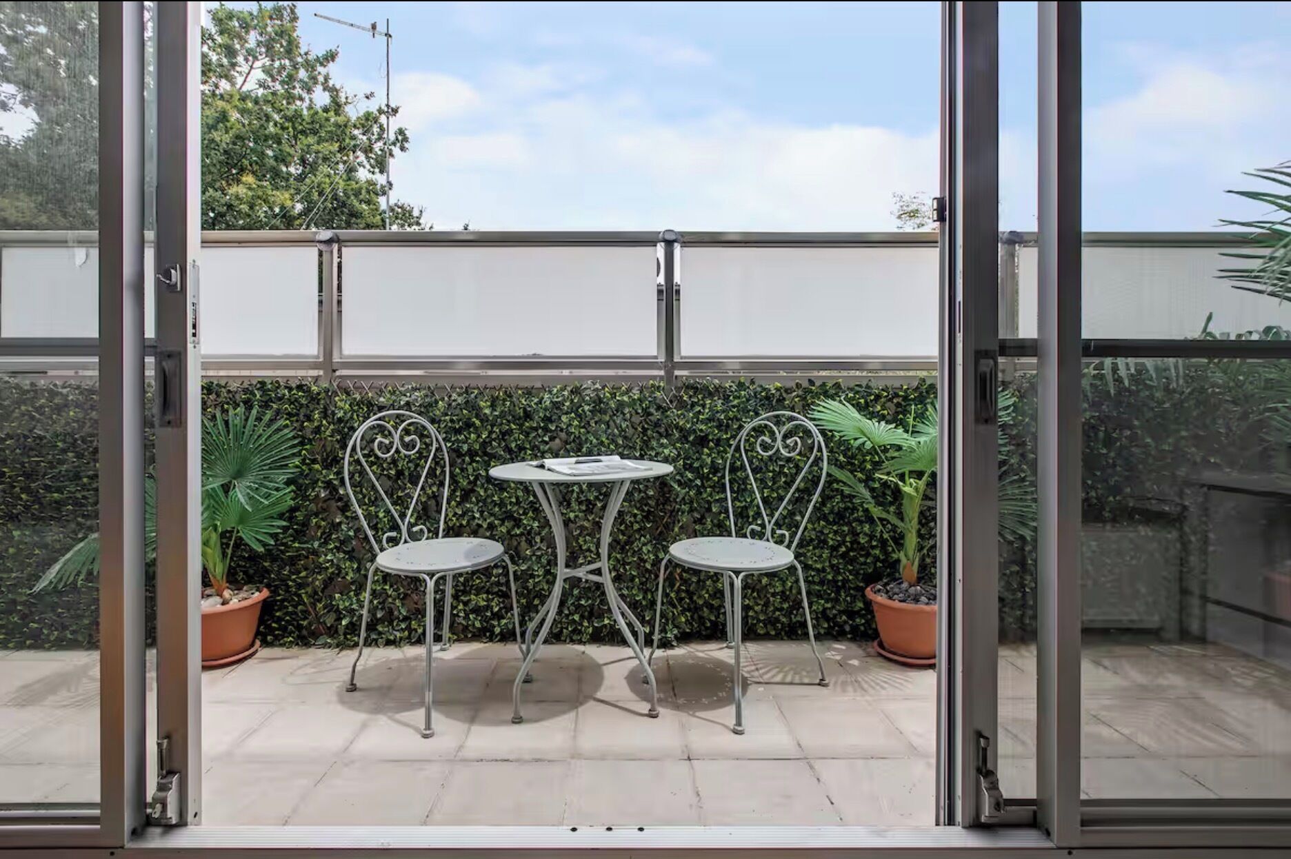 A glass-enclosed patio with two metal chairs and a small table, surrounded by potted plants and a hedge.