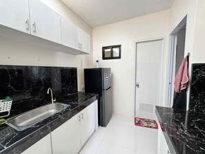 Ett kök med bänkskivor i svart marmor, en diskho i rostfritt stål, vita skåp, ett svart kylskåp och en dörr med ett ventilationsgaller.