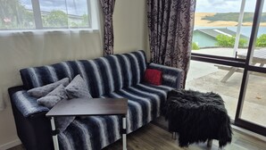 A modern living room with a striped sofa, a small table, and a view of a beach through large windows. - Studio with Hokianga Harbour & Sand Dunes view (Opononi)