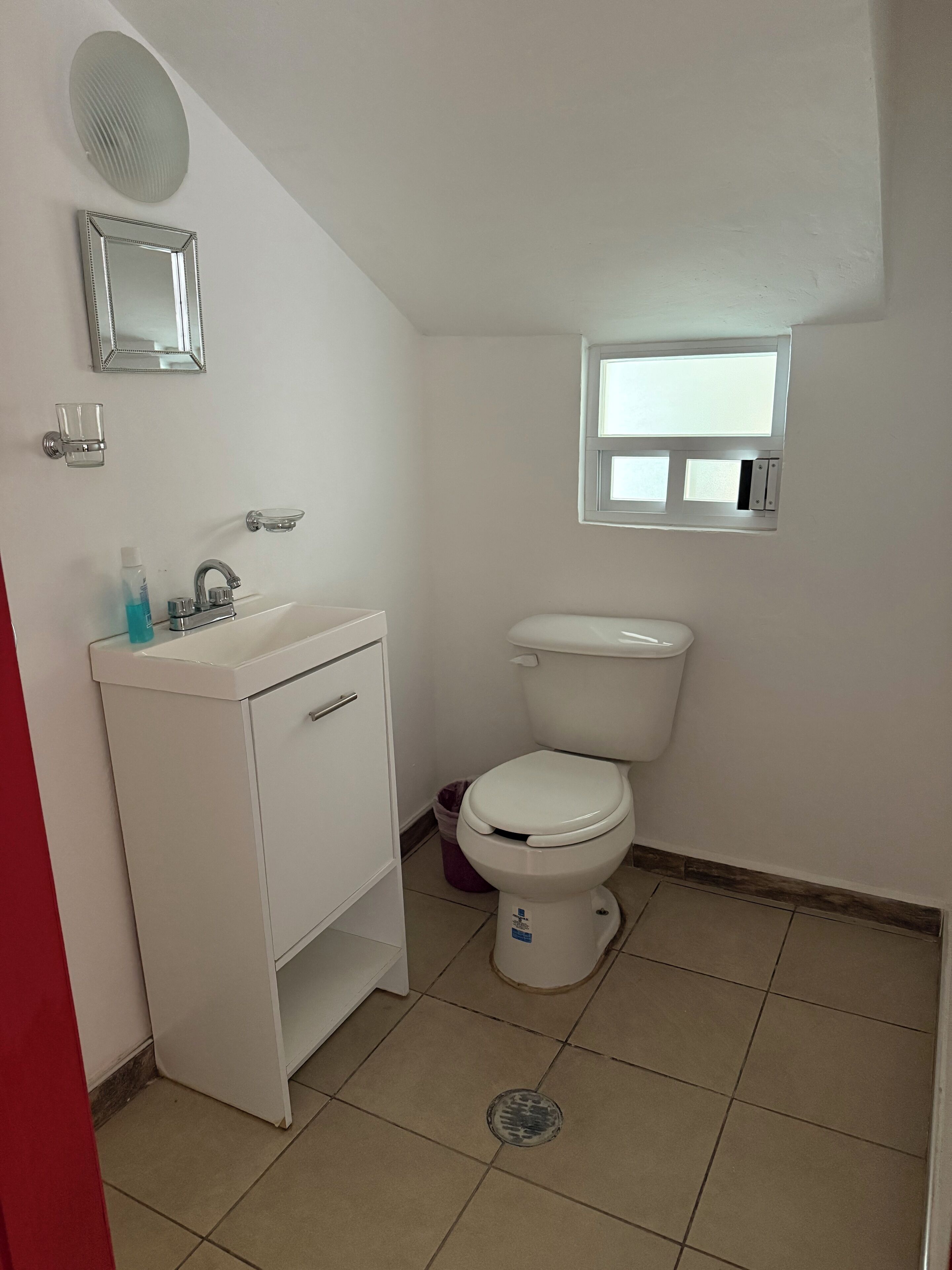 Un baño pequeño y limpio con un lavamanos, espejo y retrete blancos. Hay una ventana sobre el retrete y un estante pequeño con artículos de aseo.