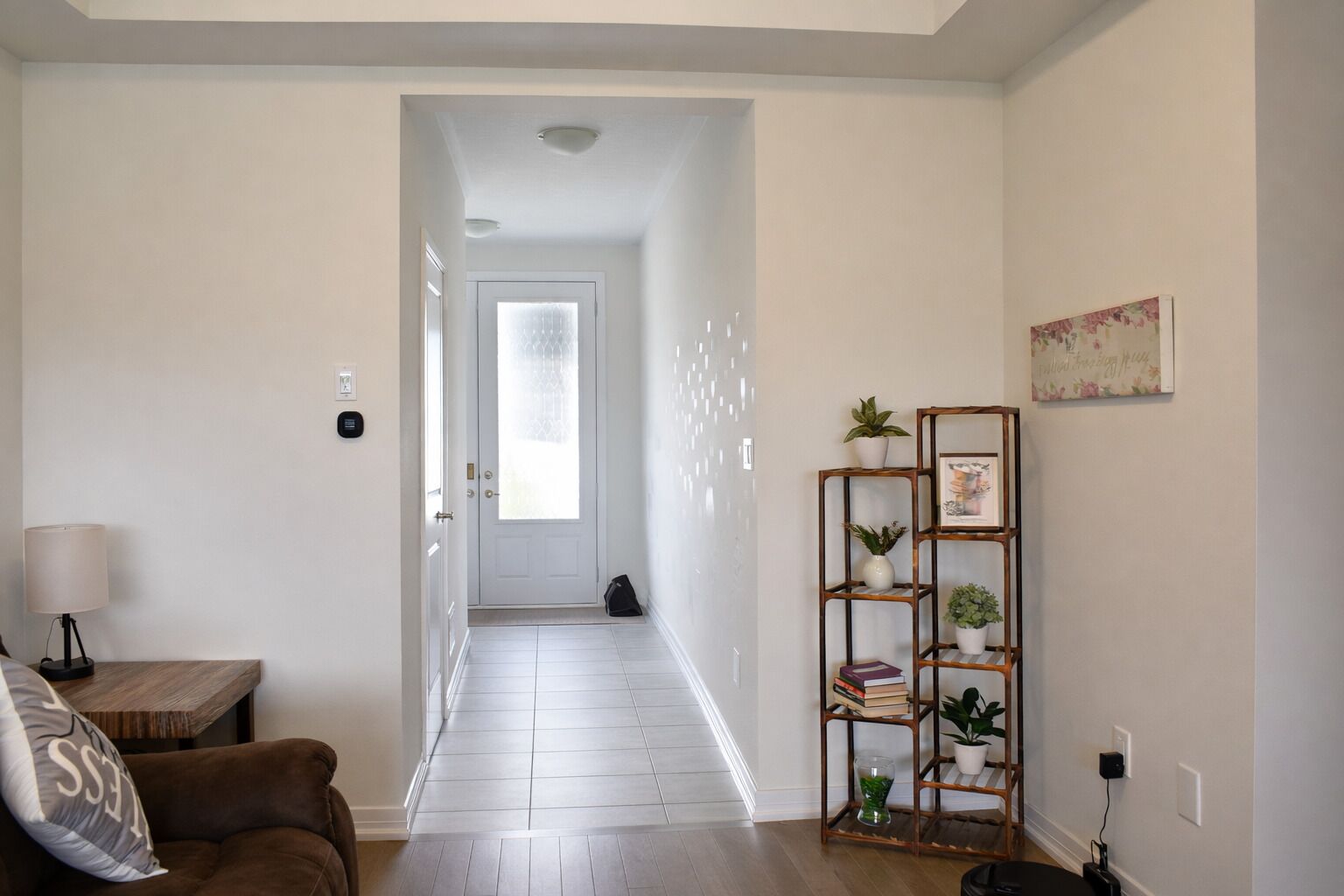 Un corridoio con una porta, un pavimento in legno, un divano marrone, una lampada, uno scaffale con piante e un quadro appeso al muro.