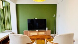Sala de estar com piso de madeira, parede verde, televisão, mesa de centro de madeira e duas poltronas brancas.