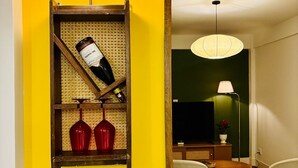 Quarto com parede amarela, estante de madeira com garrafas de vinho, televisão, mesa de jantar com cadeiras e luminária pendente.