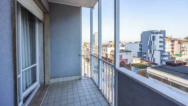 Ein Balkon mit Fliesenboden, ein Fenster mit Jalousien und Blick auf umliegende Gebäude.
