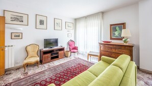 Ein Wohnzimmer mit einer Couch, einem Fernseher auf einem Holzboden, einem roten Teppich mit Muster, einem rosa Sessel und einem Holzkasten mit einer Lampe.