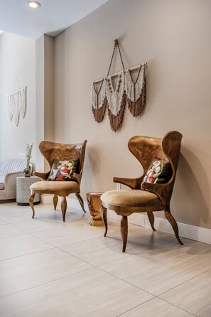 Deux fauteuils marron à revêtement en tissu, agrémentés de coussins colorés, une petite table d’appoint ronde sur laquelle est posée une plante en pot, et des décorations murales en macramé.