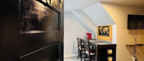 Una habitación con una puerta de madera oscura, una mesa de comedor con sillas, un televisor de pantalla plana montado en la pared y una lámpara en el techo.