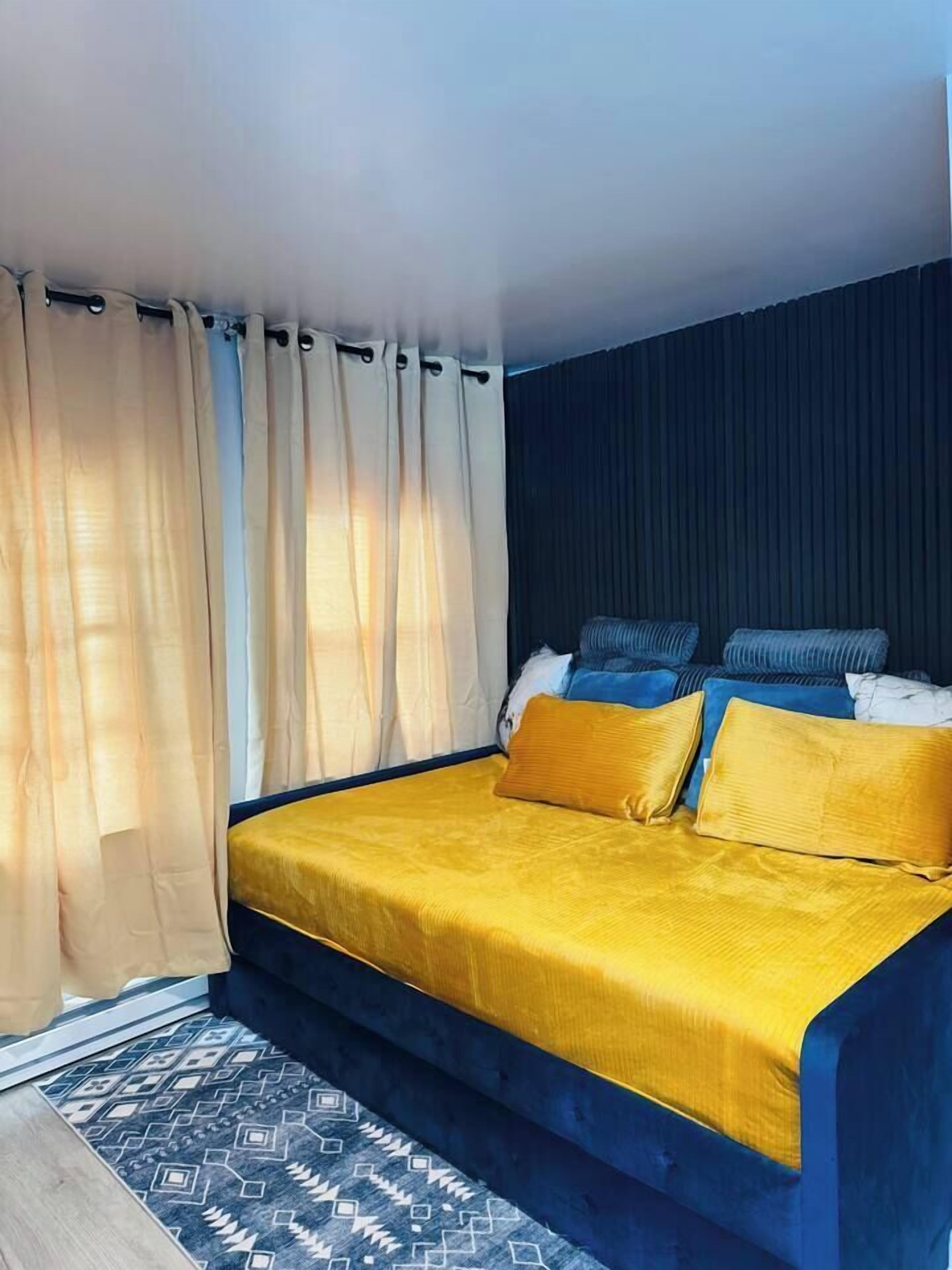 Una cama con estructura azul y colcha amarilla, cabecera oscura y cortinas de gasa.