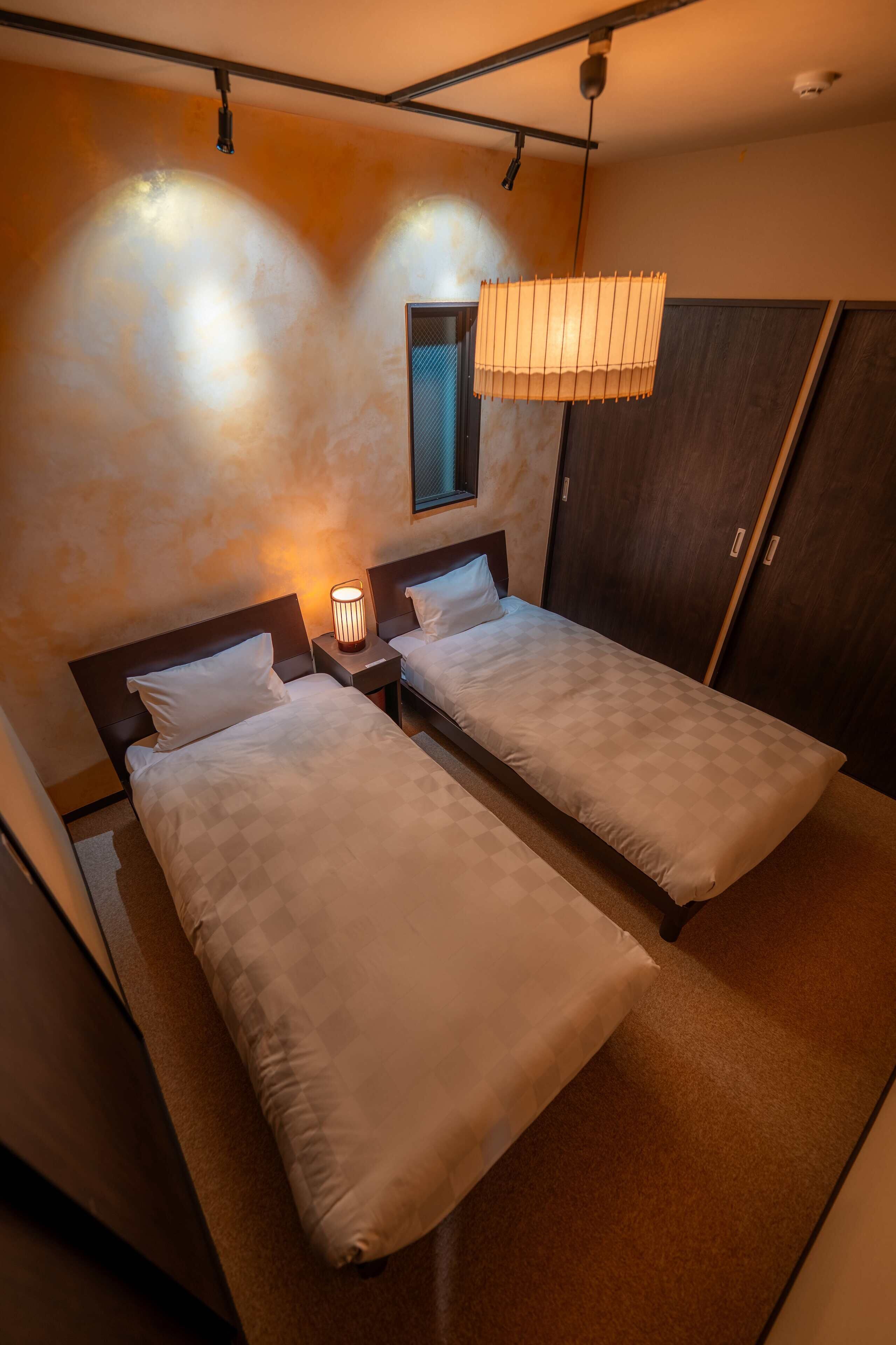 A hotel room with two single beds, a nightstand with a lamp, a wardrobe, and a window with a curtain.