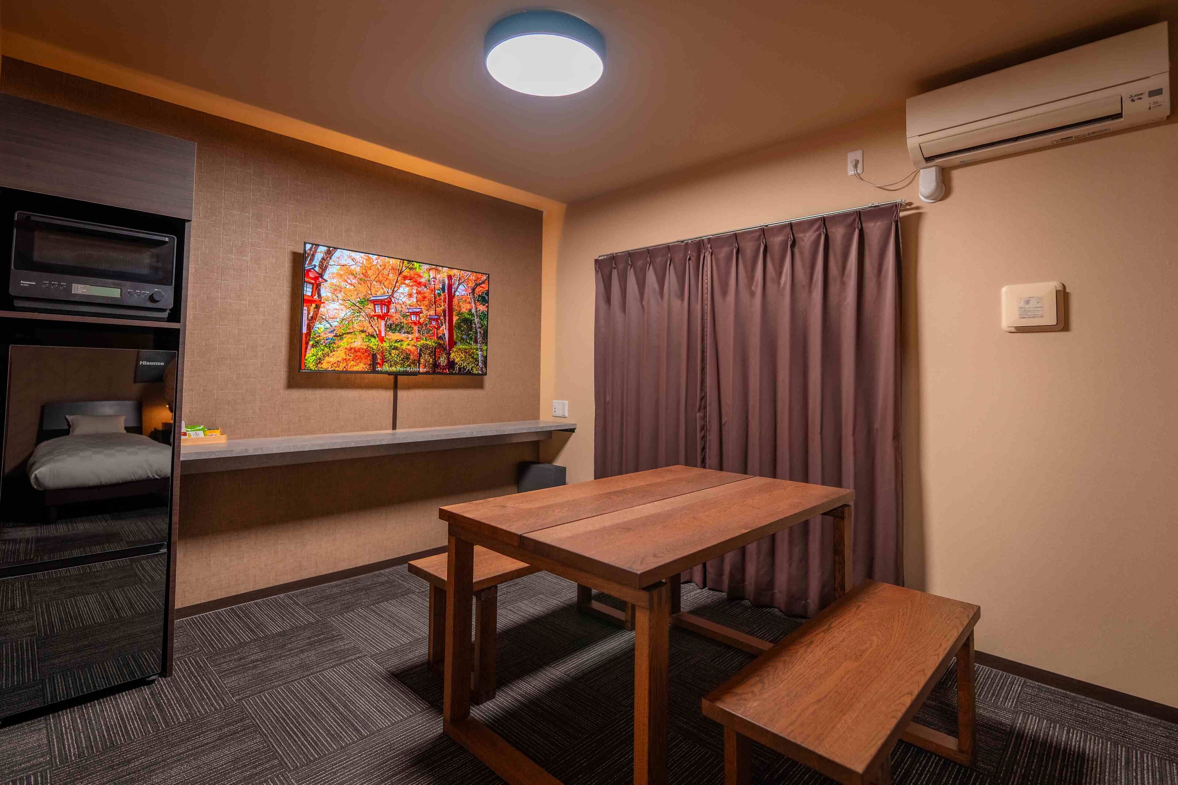 Een hotelkamer met een houten tafel, een bank, een aan de muur bevestigde televisie, een plafondlamp, een airconditioner en een raam met gordijnen.