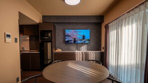 Een moderne eetruimte met een ronde tafel, stoelen, een aan de muur bevestigde televisie en een keuken op de achtergrond.