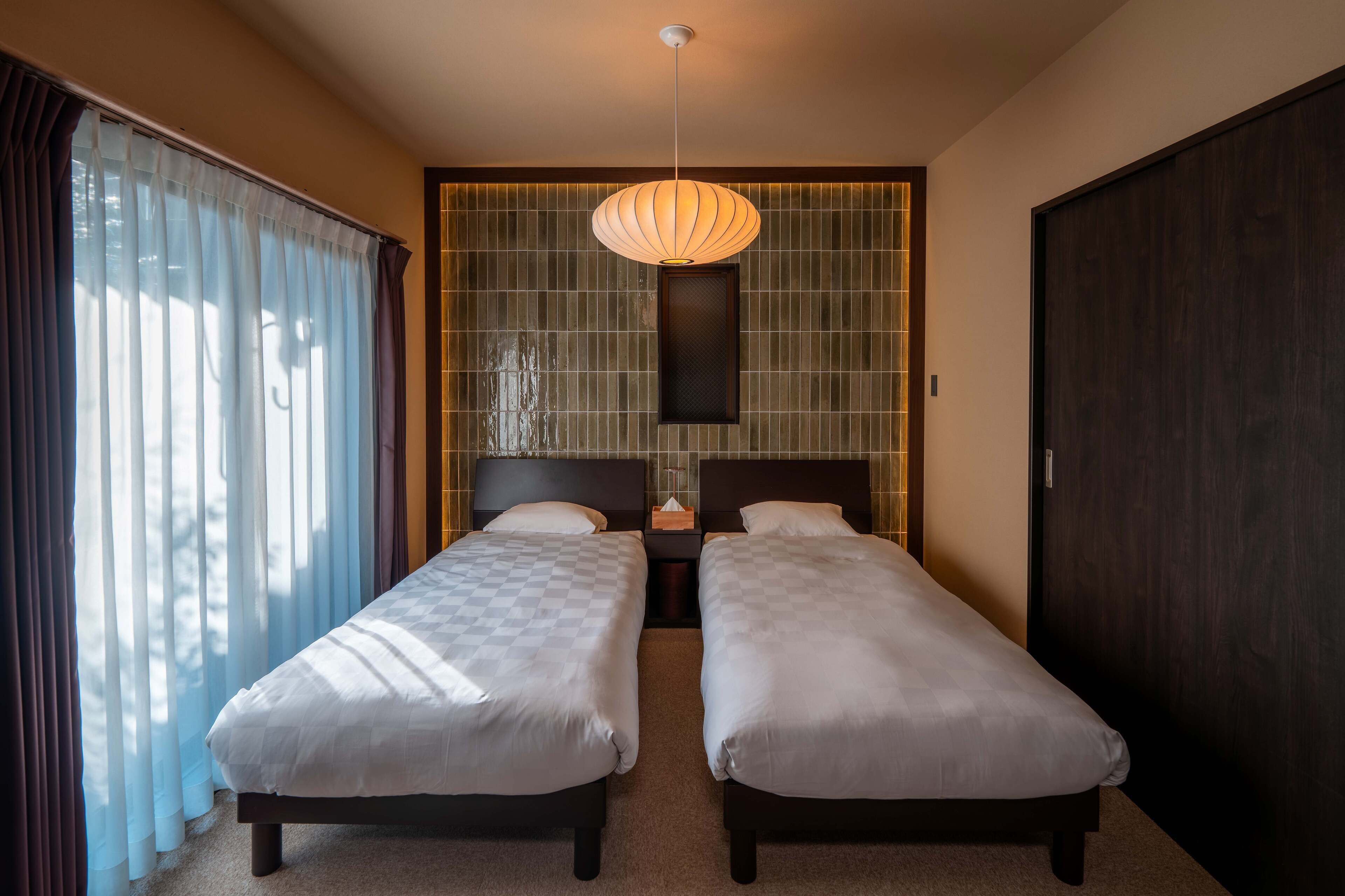 A hotel room with two single beds, a large window with curtains, a wall-mounted light fixture, and a dark wooden wardrobe.