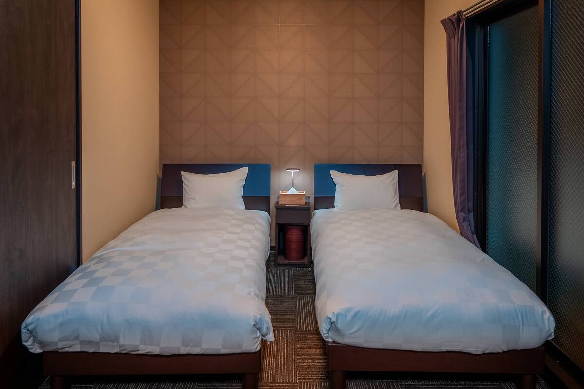 Une chambre d’hôtel avec deux lits simples, une table de chevet avec une lampe, une tête de lit en bois et une grande fenêtre avec des rideaux.
