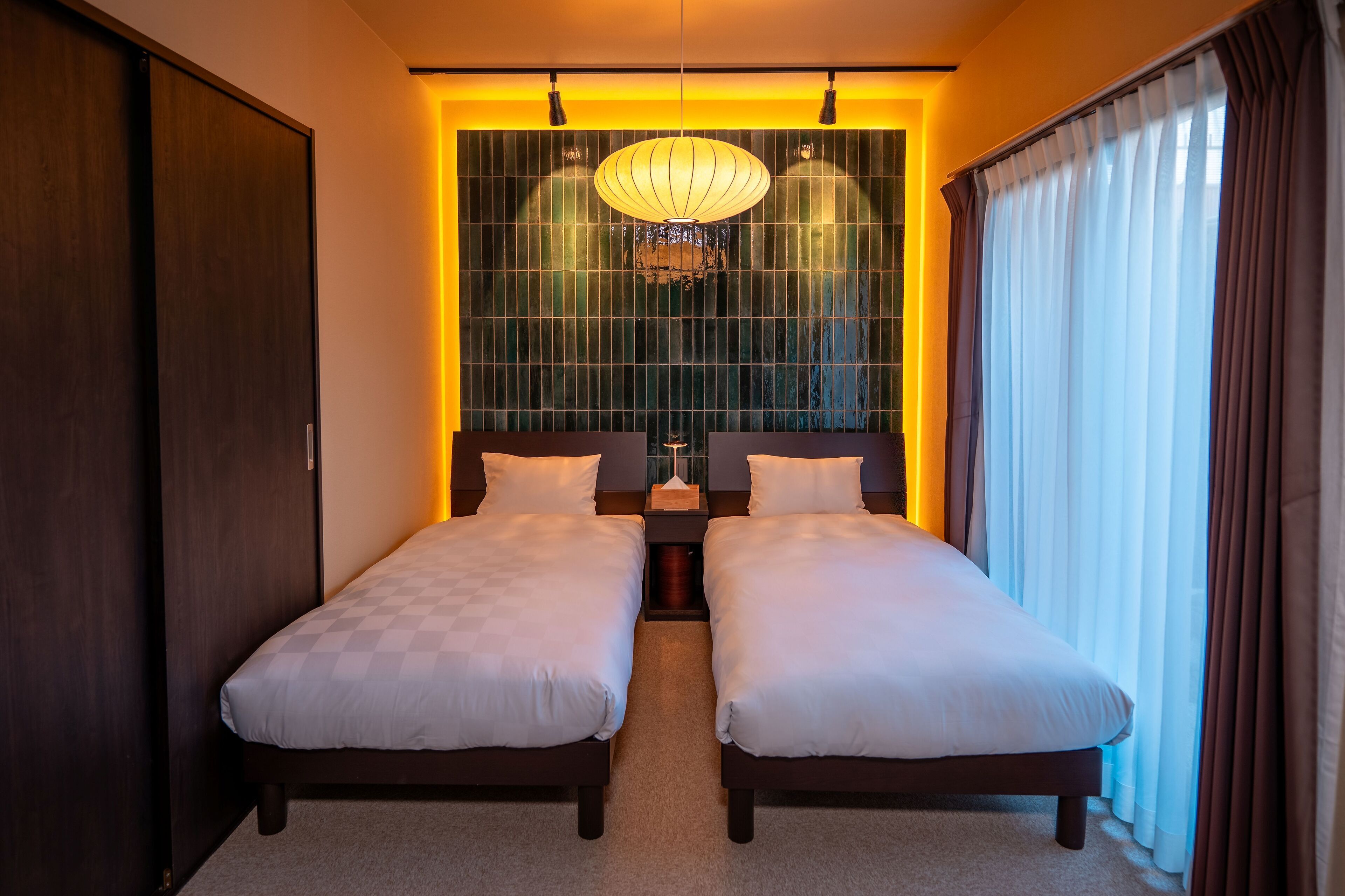 A hotel room with two single beds, a large window with curtains, a hanging light fixture, and a wardrobe.