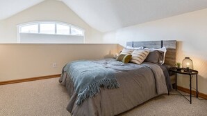 Een slaapkamer met een schuine kap, een groot raam, een bed met grijze beddengoed, een nachtkastje met een lamp, en een kleine plant.