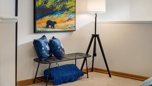 Een kamer met een schilderij van een beer in een boom, een vloerlamp, een metalen bank met kussens, en een blauwe plaid.