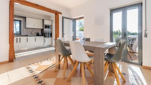 A modern kitchen with white cabinets and stainless steel appliances, a dining area with a wooden table and chairs, and a sliding glass door leading to a balcony. - Villa Sheike V3 com Piscina Privada (Tavira)