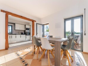 A modern kitchen with white cabinets and stainless steel appliances, a dining area with a wooden table and chairs, and a sliding glass door leading to a balcony. - Villa Sheike V3 com Piscina Privada (Tavira)