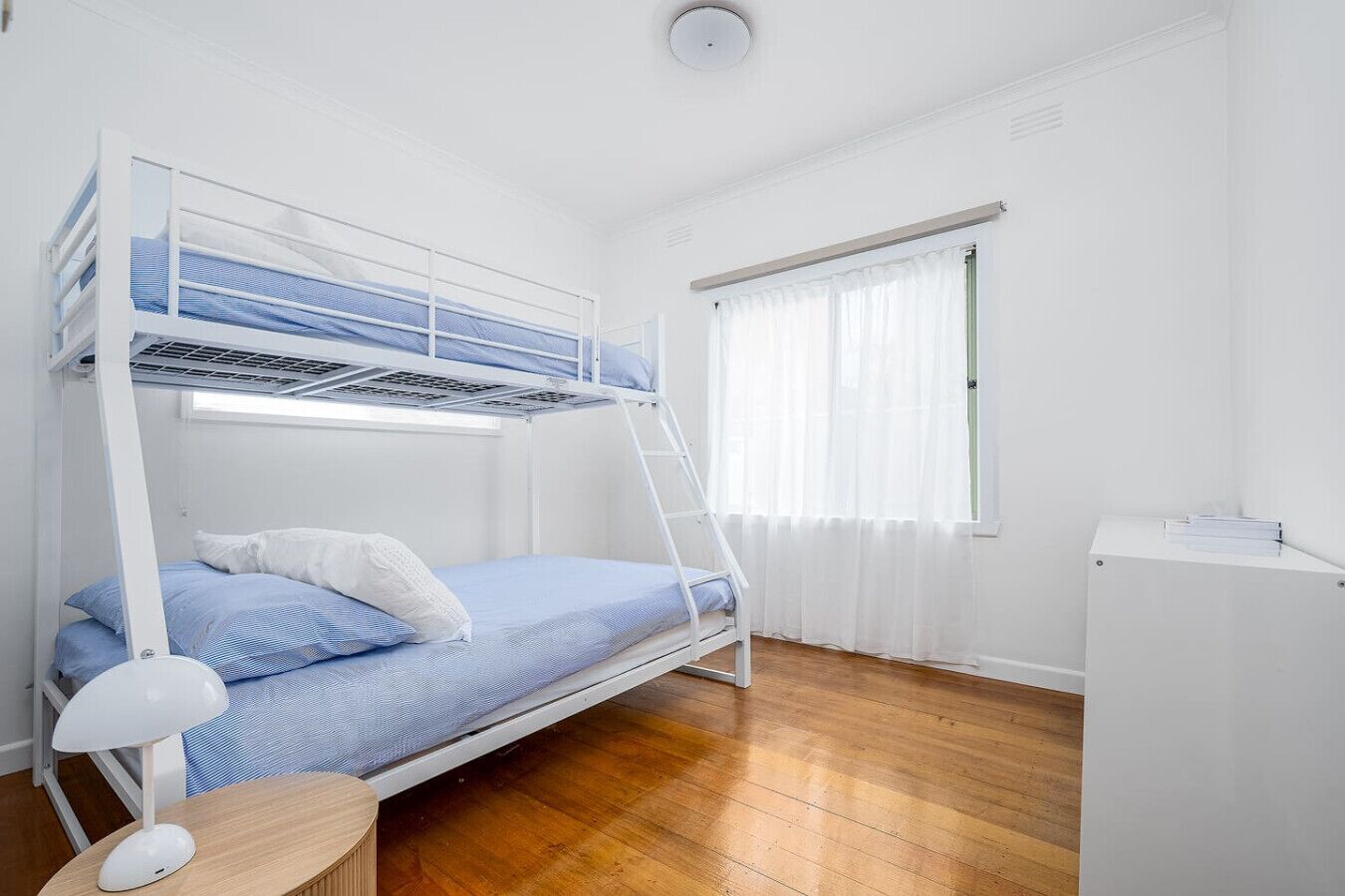 Habitación con literas, escritorio, lámpara, suelo de madera y ventana con cortinas traslúcidas.