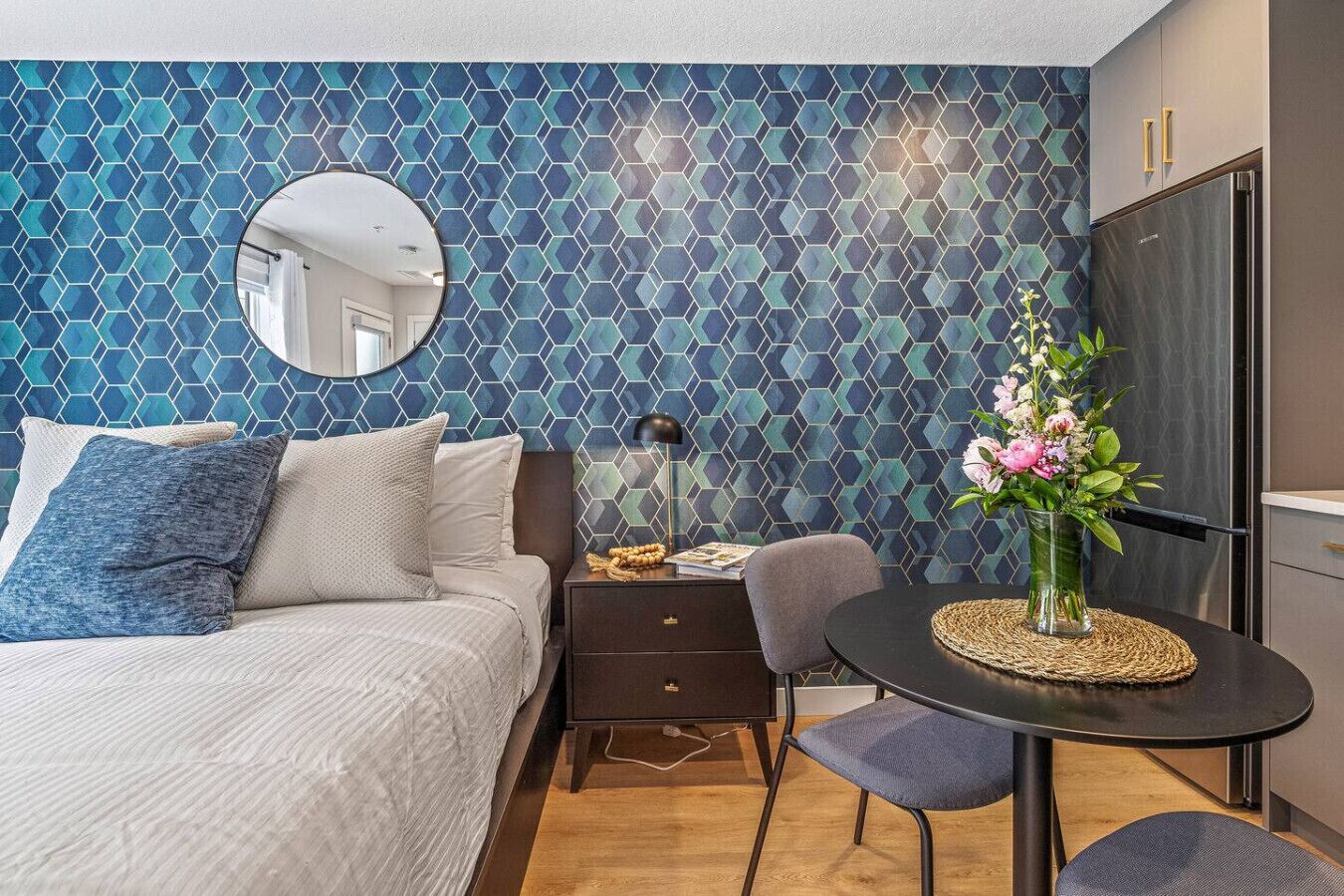 Et moderne soveværelse med væg i geometrisk mønster, et rundt spejl, en seng med blåt og hvidt sengetøj, et natbord med lampe og et lille spiseområde med bord og stole.