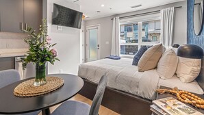 A modern hotel room with a bed, a TV, a dining table, and a view of the city through large windows. - Chick & Cozy Haven near Banff with Hot tub & Pool (Canmore)